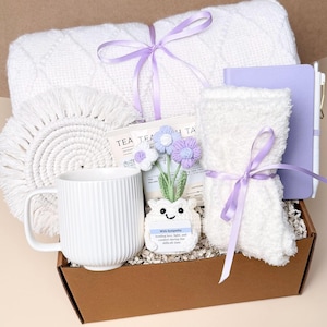 May include: A gift box filled with a white blanket, a white mug, a macrame coaster, a small notebook, a pair of fuzzy socks, and a small decorative flower pot. The items are tied with lavender ribbons. The box is labeled "Box #8".