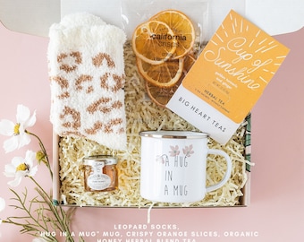 Op de afbeelding: Een cadeaudoos gevuld met knusse items voor een ontspannen avond thuis. Het bevat luipaardprint sokken, een mok met de tekst "A Hug in a Mug", gedroogde sinaasappelschijfjes, een potje honing en een theezakje met de tekst "Cup of Sunshine" met de tekst "Golden Turmeric and Ginger" en "Herbal Tea".