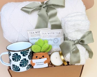 May include: A gift box containing a white blanket, a mug with a monstera leaf design, two tea packets, a small candle, and a crocheted cactus holding a sign that reads "Life would succ without you!" The box is tied with sage green ribbons.