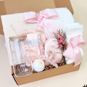 May include: A brown gift box filled with a variety of self-care items, including a white blanket, a pink eye mask, a glass tumbler with a straw, a bath bomb, a pink face mask, a bottle of bath salts, and a bouquet of pink flowers.