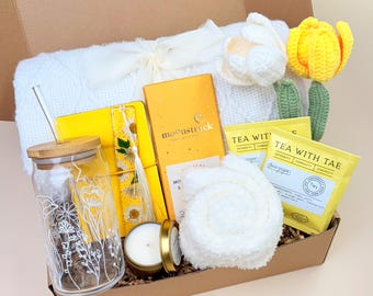 May include: A yellow and white gift box filled with various items, including a white knitted blanket, a yellow journal, a glass jar with a straw, a yellow and white bookmark, a yellow chocolate bar, a white hand towel, a yellow candle, and two tea bags. The box is labeled "Tea With Tae" and the chocolate bar is labeled "Moonstruck".