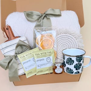 K&ouml;nnte beinhalten: Geschenkbox gef&uuml;llt mit einer wei&szlig;en Decke, einem Holzschneidebrett, einem gestreiften Handtuch, Teebeuteln, getrockneten Orangenscheiben, einem Untersetzer, einer Tasse mit gr&uuml;nem Bl&auml;ttermuster und einem Honigglas. Die Box ist mit gr&uuml;nen B&auml;ndern gebunden.