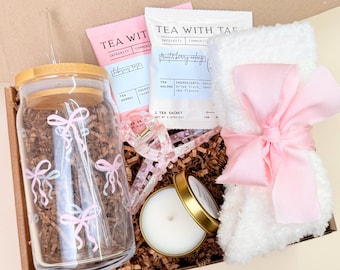 May include: A gift box filled with a glass jar with pink bow designs, tea sachets, a white fluffy item tied with a pink ribbon, a candle, and a hair clip. The jar has a bamboo lid and a straw. The box is filled with brown paper.