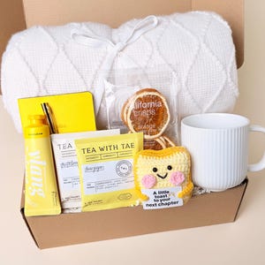 May include: A gift box containing a white knit blanket, a yellow notebook with a gold pen, a tube of yellow hand cream, a packet of Tea With Tae lemon ginger tea, a bag of California Crisps orange slices, and a white ribbed mug with a crochet toast.