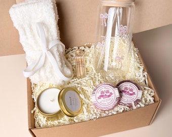 Op de afbeelding: Een geschenkdoos met een paar witte pluizige sokken vastgebonden met een lint, een doorzichtige glazen beker met bamboe deksel, een glazen rietje, roze strikjes, een flesje lucifers, een goudkleurig kaarsblik en twee ronde douche-bruistabletten.