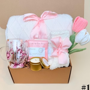 Pode incluir: Uma caixa de presente com um cobertor branco, uma caneca de vidro com detalhes florais, saquinhos de chá, uma vela e um pequeno item de pelúcia, todos amarrados com fitas rosa. Tulipas de crochê em rosa e branco adicionam decoração.