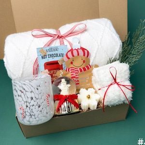 May include: A gift basket filled with winter-themed items. Includes a white blanket, a glass with candy cane accents, a snow globe, hot chocolate mix, a gingerbread man ornament, a white towel, and a snowflake-shaped candle. All items are arranged in a brown box.