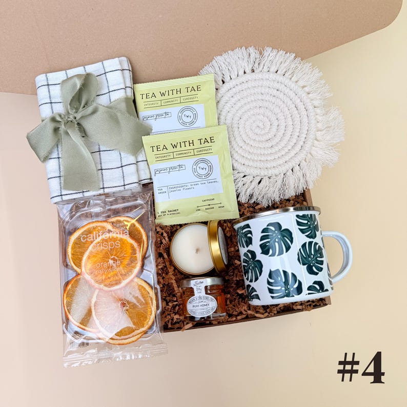 K&ouml;nnte beinhalten: Eine Geschenkbox mit einem wei&szlig;-gr&uuml;nen karierten Tuch, das mit einem gr&uuml;nen Band gebunden ist, Teebeuteln, einem wei&szlig;en Makramee-Untersetzer, einer Kerze, einem Honigglas, einer Tasse mit gr&uuml;nem Bl&auml;ttermuster und Orangenscheiben.