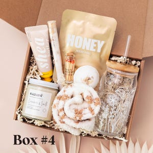 May include: A gift box containing a tube of shea butter, a glass tube of bath salts, a gold pouch labeled HONEY NOURISHING, a small bottle of matches, a bath bomb, a candle labeled SUCCULENT, a fuzzy tan and white sock, and a glass cup with a bamboo lid.