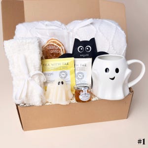 May include: A gift box containing a white knit blanket, a white fuzzy towel with a ribbon, a bag of California Crisps, a black bat-shaped face mask, two Tea With Tae tea packets, a ghost-shaped candle, a jar of honey, and a white ghost mug.