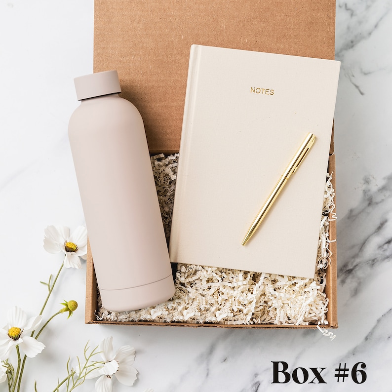 May include: A brown cardboard box with a light brown notebook, a gold pen, and a light pink water bottle inside. The box is filled with shredded paper. The text "NOTES" is printed on the front of the notebook.