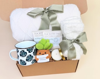 May include: A gift box containing a white knit blanket tied with a sage green ribbon, a white enamel mug with green monstera leaf prints, a crocheted succulent in a pot with a sign reading Life would succ without you, tea bags, and a candle.