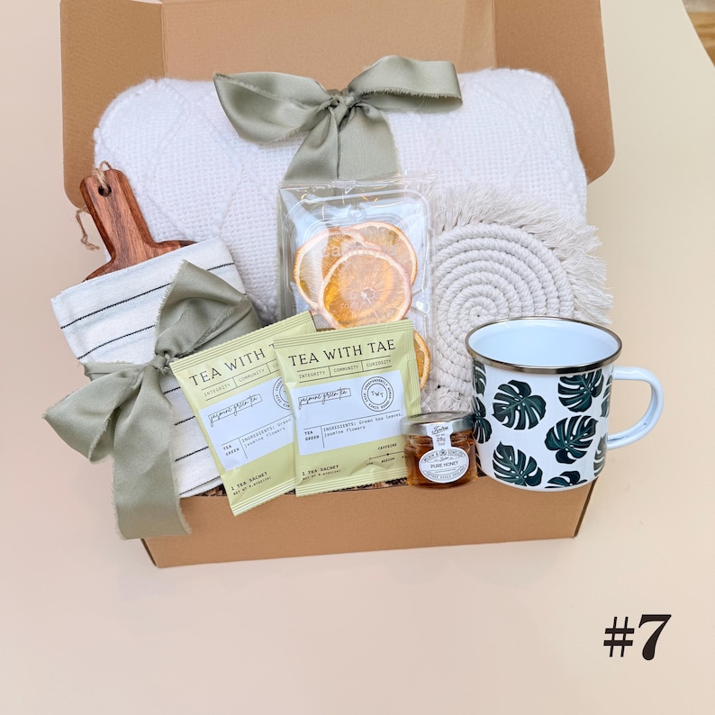 K&ouml;nnte beinhalten: Geschenkbox gef&uuml;llt mit einer wei&szlig;en Decke, einem Holzschneidebrett, einem gestreiften Handtuch, Teebeuteln, getrockneten Orangenscheiben, einem Untersetzer, einer Tasse mit gr&uuml;nem Bl&auml;ttermuster und einem Honigglas. Die Box ist mit gr&uuml;nen B&auml;ndern gebunden.