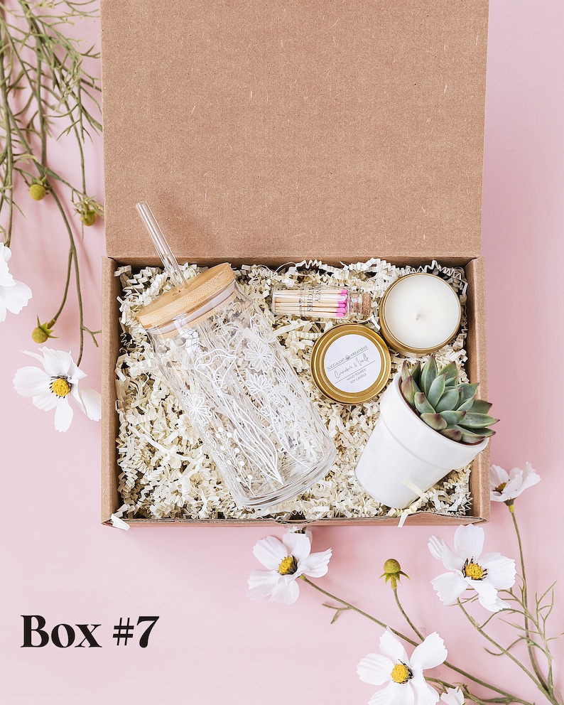 May include: A brown cardboard box filled with a gift set. The box contains a glass tumbler with a straw, a small candle, a small plant in a white pot, and a small glass bottle with matches. The box is surrounded by white flowers.