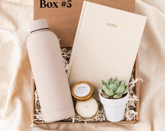 May include: A cardboard gift box labeled Box #5 containing a matte light pink water bottle, a cream-colored notebook with the word NOTES printed in gold, a small candle in a gold tin, and a green succulent in a white ceramic pot.