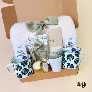 K&ouml;nnte beinhalten: Geschenkbox gef&uuml;llt mit einer wei&szlig;en Decke, zwei Tassen mit gr&uuml;nen Bl&auml;ttermustern, Kaffee, einer Kerze, einer Sukkulente und einer Schokoladentafel. Die Box ist mit einer gr&uuml;nen Schleife gebunden. Die Nummer 9 befindet sich unten rechts.