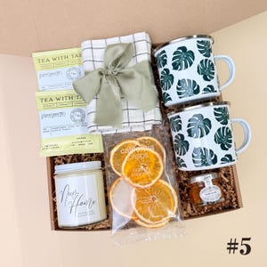 K&ouml;nnte beinhalten: Eine Geschenkbox mit zwei wei&szlig;en Emaille-Tassen mit gr&uuml;nen Blattmustern, einer Kerze mit der Aufschrift "New Home", Teesachets, getrockneten Orangenscheiben, einem kleinen Glas Honig und einem karierten Tuch, das mit einem gr&uuml;nen Band gebunden ist.