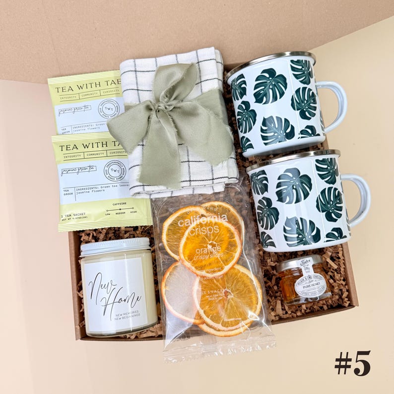 K&ouml;nnte beinhalten: Eine Geschenkbox mit zwei wei&szlig;en Emaille-Tassen mit gr&uuml;nen Blattmustern, einer Kerze mit der Aufschrift "New Home", Teesachets, getrockneten Orangenscheiben, einem kleinen Glas Honig und einem karierten Tuch, das mit einem gr&uuml;nen Band gebunden ist.