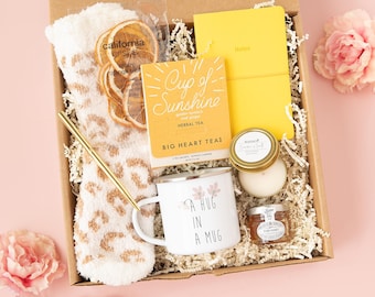 May include: A gift box containing a pair of fuzzy leopard print socks, a package of California Crisps, a yellow notebook, a box of Big Heart Tea labeled Cup of Sunshine, a white mug with the text A HUG IN A MUG, a candle, and honey.