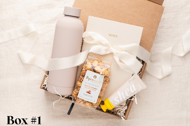 May include: A gift box filled with a pink water bottle, a notebook with the word "NOTES" on the cover, a bag of caramel popcorn, and a tube of shea butter lotion.