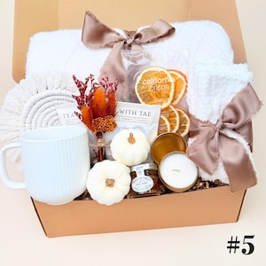 May include: A gift box containing a white ribbed mug, a white textured blanket tied with a mauve ribbon, a white towel with a mauve ribbon, two small white pumpkins, a jar of honey, dried orange slices, a tea packet, and a dried floral bouquet.
