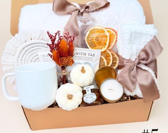 May include: A gift box containing a white ribbed mug, a white textured blanket tied with a mauve ribbon, a white towel with a mauve ribbon, two small white pumpkins, a jar of honey, dried orange slices, a tea packet, and a dried floral bouquet.