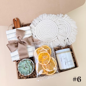K&ouml;nnte beinhalten: Eine Geschenkbox mit verschiedenen Artikeln. Enth&auml;lt ein kleines Schneidebrett mit gestreiftem Handtuch und Schleife, eine Sukkulente, getrocknete Orangenscheiben, zwei wei&szlig;e Makramee-Untersetzer und eine Kerze mit der Aufschrift "Nur Home".