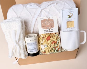Peut inclure: Un coffret cadeau contenant une couverture blanche en tricot torsadé nouée d'un ruban, une tasse blanche texturée, un sachet de soupe Cooke Tavern Yankee Doodle Chicken Noodle, des crackers Jocelyn & Co. Olive Oil & Sea Salt et une boîte de thé.
