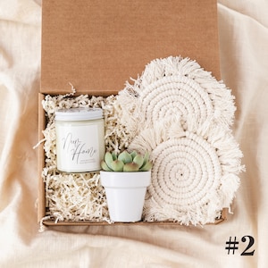 K&ouml;nnte beinhalten: Geschenkbox mit einer Kerze, einer kleinen Sukkulente in einem wei&szlig;en Topf und zwei cremefarbenen Makramee-Untersetzern. Die Kerze hat ein wei&szlig;es Etikett mit dem Schriftzug "Our Home". Die Box ist mit Papier gef&uuml;llt.