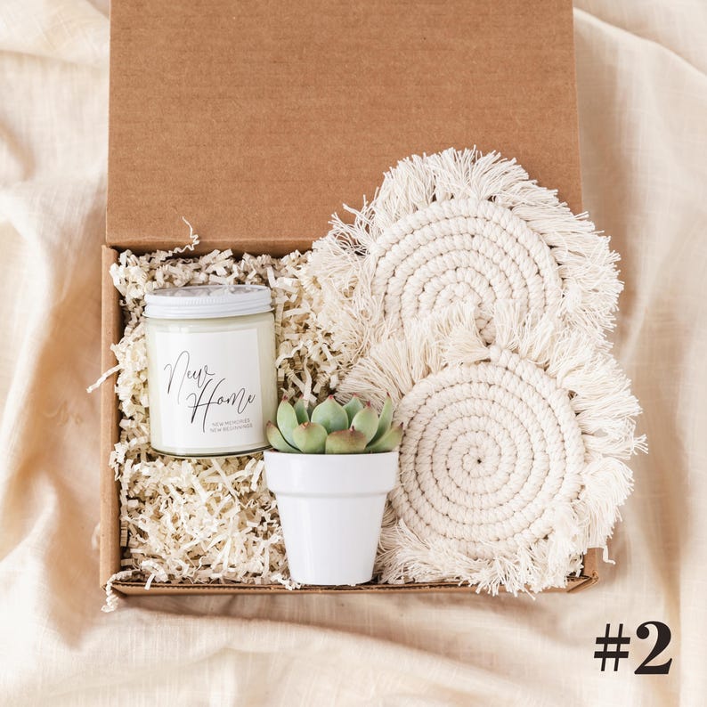 K&ouml;nnte beinhalten: Geschenkbox mit einer Kerze, einer kleinen Sukkulente in einem wei&szlig;en Topf und zwei cremefarbenen Makramee-Untersetzern. Die Kerze hat ein wei&szlig;es Etikett mit dem Schriftzug "Our Home". Die Box ist mit Papier gef&uuml;llt.