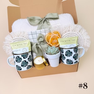 K&ouml;nnte beinhalten: Eine Geschenkbox mit einer wei&szlig;en Decke, zwei Tassen mit gr&uuml;nen Monstera-Blattmustern, Teebeuteln, getrockneten Orangenscheiben, einer kleinen Sukkulente, einer Kerze und einer dekorativen Schleife. Die Box ist mit der Nummer 8 versehen.