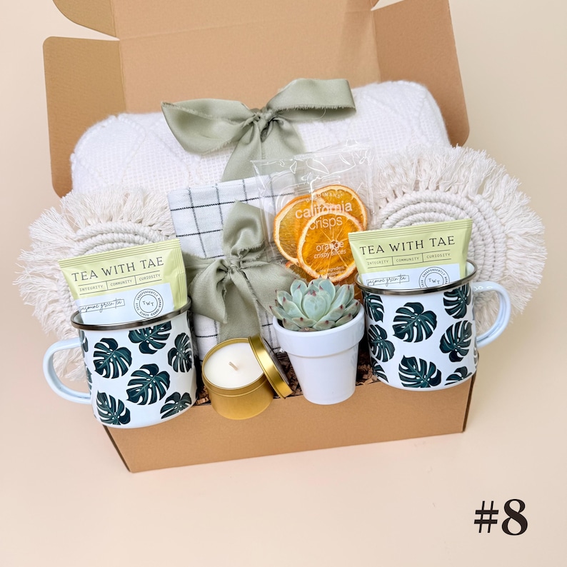 K&ouml;nnte beinhalten: Eine Geschenkbox mit einer wei&szlig;en Decke, zwei Tassen mit gr&uuml;nen Monstera-Blattmustern, Teebeuteln, getrockneten Orangenscheiben, einer kleinen Sukkulente, einer Kerze und einer dekorativen Schleife. Die Box ist mit der Nummer 8 versehen.