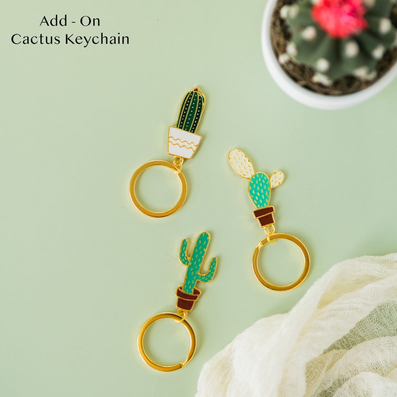 Op de afbeelding: Drie gouden cactus sleutelhangers met groene en bruine details. Elke hanger heeft een ander cactus ontwerp en is bevestigd aan een gouden sleutelring.
