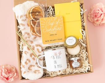 May include: A gift box containing a pair of fuzzy white socks with a tan leopard print, a package of California Crisps, a yellow notebook, a box of Big Heart Tea, a gold pen, a white mug with floral art and text, a candle, and honey.