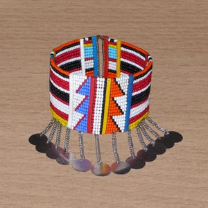 Può includere: Un braccialetto largo di perline con un design colorato. Il braccialetto presenta perline rosse, nere, bianche, blu e gialle. Piccoli dischi d'argento pendono dal basso, attaccati da catene d'argento.