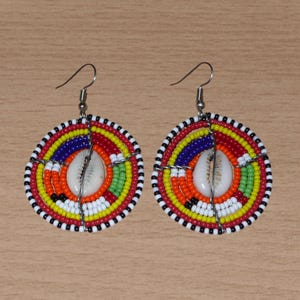 Peut inclure: Une paire de boucles d'oreilles perlées faites à la main. Chaque boucle d'oreille présente un motif circulaire avec des anneaux concentriques de perles rouges, jaunes, oranges, vertes, bleues et blanches. Un coquillage blanc est centré dans chaque boucle d'oreille.
