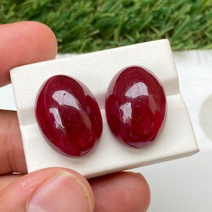 May include: Two oval-shaped, deep red gemstones on a white display stand.