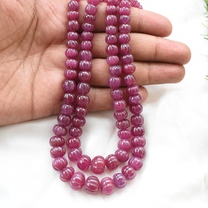 May include: A strand of pink ruby beads, each bead is carved with a ribbed pattern. The beads are strung on a red cord.