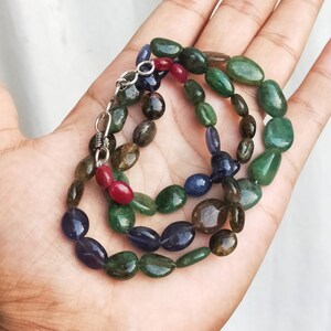 May include: A multi-colored gemstone necklace with green, blue, and red stones. The necklace is made of oval-shaped beads and has a silver clasp.