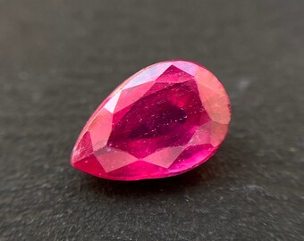 Puede incluir: Una piedra preciosa de color rojo intenso en forma de pera con un corte facetado. La piedra preciosa se coloca sobre un fondo gris oscuro.