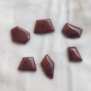 May include: Six irregularly shaped, deep red gemstones on a white fabric background.