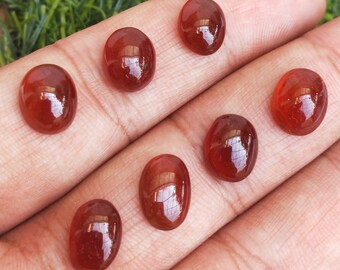 May include: Seven oval-shaped, translucent, red gemstones with a glossy finish. The gemstones are arranged in a row on a hand.
