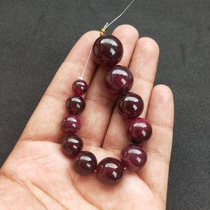 Könnte beinhalten: Eine Reihe dunkelroter Edelsteinperlen, auf einem Silberdraht aufgefädelt. Die Perlen sind rund und glatt und in abgestufter Größe angeordnet, wobei die größte Perle oben liegt.