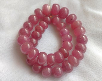 May include: A strand of pink gemstone beads, likely ruby, strung on a clear thread. The beads are round and smooth, with a slight variation in size. The beads are arranged in a circular pattern, with two strands overlapping.