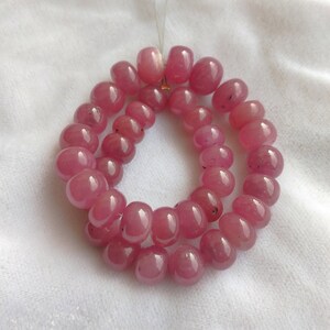 May include: A strand of pink gemstone beads, likely ruby, strung on a clear thread. The beads are round and smooth, with a slight variation in size. The beads are arranged in a circular pattern, with two strands overlapping.