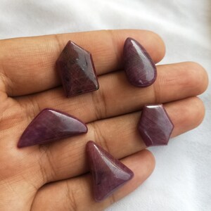 Könnte beinhalten: Fünf violette Edelstein-Cabochons in verschiedenen Formen, darunter eine Träne, ein Hexagon und ein Dreieck.