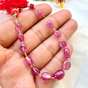 Peut inclure: Un brin de perles de pierres précieuses ovales roses avec un enfileur en fil d'or. Les perles sont enfilées sur un fil d'or et ont un pompon rouge à une extrémité.