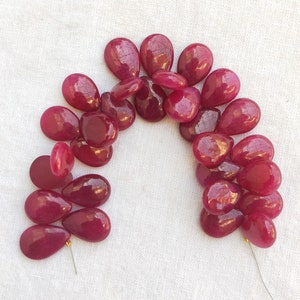 May include: A strand of deep red gemstone beads in a teardrop shape. The beads are strung on a thin wire and are arranged in a semi-circle.
