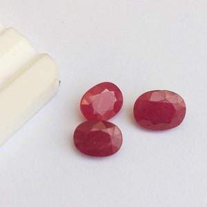 以下が含まれることがあります： 白い表面に置かれた3つの楕円形のルビーの宝石。宝石は濃い赤色で、ファセットカットされています。