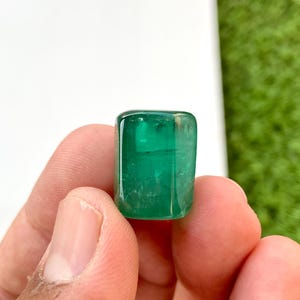 May include: A vibrant green emerald gemstone, cut into a rectangular shape, is held in a hand. The gemstone is smooth and polished, with a slight sheen.
